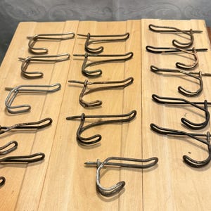 May include: A collection of metal hooks with attached screws, arranged on a light-colored wooden surface. The hooks are various shades of silver and dark brown, with a curved design for hanging items.