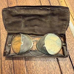 May include: Antique spectacles with wire frames and mesh lenses, housed in a brown velvet-lined case. The lenses have a yellowed appearance, and the case is open, revealing the glasses. The glasses are resting on a wooden surface.