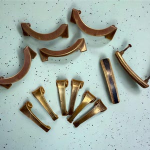 May include: An assortment of vintage brass cabinet hardware, including curved handles and smaller pulls. The hardware has a warm, aged patina, with some pieces featuring a dark blue or black accent. The pieces are arranged on a light blue surface.