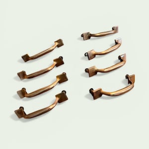 May include: A collection of ten antique bronze-colored cabinet handles. Each handle has a curved bar with square mounting plates on each end. The handles are arranged on a light background.