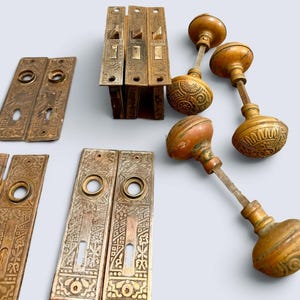 May include: A collection of antique brass door hardware including three rectangular backplates with intricate embossed patterns, two mortise lock mechanisms, and three round door knobs with matching decorative engravings and metal spindles.