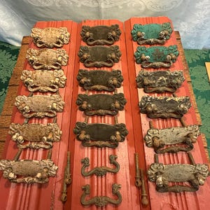 May include: A collection of antique drawer pulls and handles in various styles and finishes. The pulls are mounted on red wooden boards. Finishes include aged white, dark bronze, and turquoise. Some pulls have ornate designs.