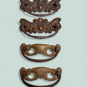 May include: Four antique bronze drawer pulls with ornate designs. Two pulls have a detailed, curved shape with decorative elements, while the other two have a simpler, circular design with a curved handle. The pulls are arranged vertically against a light blue background.