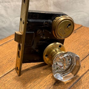 May include: An antique door lock set with a black metal body, brass faceplate, and a clear, faceted glass doorknob. The lock mechanism includes a brass Yale cylinder. The set is designed for a vintage door.