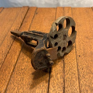 May include: An antique metal tool with a circular, multi-holed cutting head and a black handle. The tool has a worn, aged appearance, suggesting it's vintage. The circular head has numbers etched around the holes.