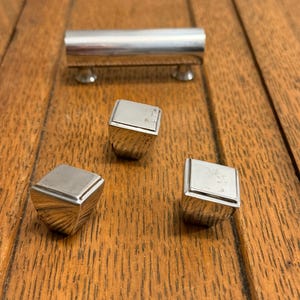 May include: A set of silver-toned cabinet hardware, including a long, cylindrical handle and three square-topped knobs. The hardware is set against a backdrop of a wooden surface with visible grain.