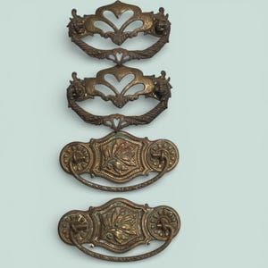May include: Four antique brass drawer pulls. Two pulls have a heart-shaped design with ornate details and lion head accents. The other two have a rectangular shape with a leaf design and circular handles. All are mounted on a light blue background.