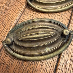 May include: An oval-shaped brass drawer pull with a weathered, aged patina. The hardware features a central raised oval element surrounded by concentric rings and a hinged, curved drop handle attached to two small circular mounting points on the sides.