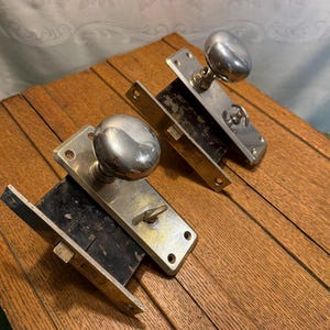 May include: Two vintage mortise door locks with polished silver-tone round knobs and rectangular metal faceplates. Each lock assembly features a black metal box housing, a protruding latch bolt, and a small integrated thumb turn mechanism.