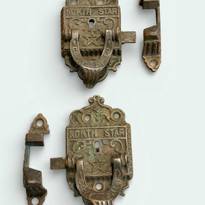 May include: Two antique metal latches with the text "NORTH STAR" and a star emblem. The latches are ornate with a horseshoe-shaped handle and a keyhole. The metal has a tarnished, aged appearance.