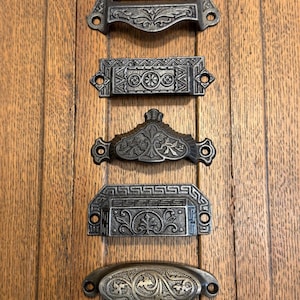 May include: Five antique drawer pulls with ornate, detailed designs in a dark, metallic finish. The pulls are arranged vertically against a wooden background, showcasing various shapes and patterns, including floral and geometric motifs.