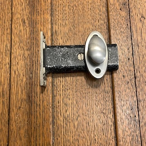 May include: A black metal door latch with a silver oval handle and a rectangular base. The latch is mounted on a wooden surface with visible wood grain.