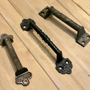 May include: Three antique-style door handles in bronze and black. The handles have ornate designs, including floral patterns and decorative end pieces. They are arranged on a light wood surface.