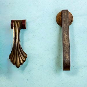 May include: Two antique brass drawer pulls. One features a fan-shaped design with a red accent, while the other is a simple rectangular pull with a circular base. Both have a distressed finish and are set against a light blue background.
