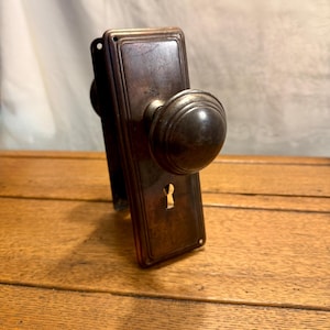 May include: Antique bronze door knob set with a rectangular backplate and a round, ridged knob. The keyhole is visible on the backplate. The hardware is set against a wooden surface.