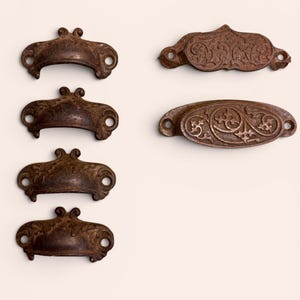 May include: A collection of antique bronze drawer pulls. The pulls have ornate floral designs and a weathered finish. The hardware includes both curved and oval shapes, each with mounting holes for easy installation. Ideal for vintage furniture restoration.
