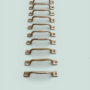 May include: A collection of nine vintage-style brass drawer pulls arranged vertically. Each pull has a curved handle and two mounting holes. The brass has a slightly aged, tarnished appearance, suggesting antique or rustic charm.