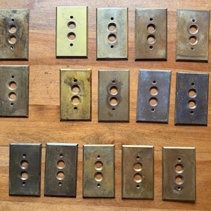 May include: A collection of rectangular, vintage brass electrical switch plates. Each plate has two or three circular holes for switches and screw holes. The plates vary in color from gold to dark brown, showing signs of age and wear.