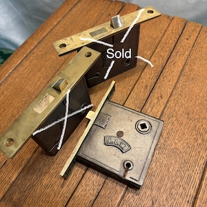 May include: Three rectangular mortise locks with dark brown metal bodies and brass faceplates. Each lock features a square spindle hole and a protruding latch bolt. One lock is marked with the letters B.L.W. Two locks have white markings.