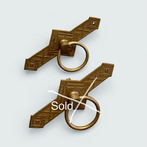 May include: Two antique brass door pulls with ring handles. The pulls have a geometric design and are marked "Sold". The hardware is likely for a cabinet or drawer.