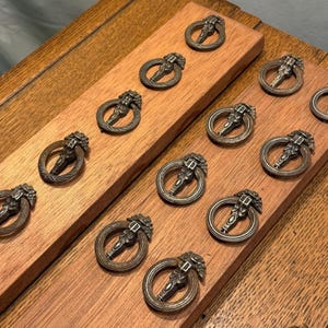 May include: A collection of twelve circular metal drawer pulls with a dark bronze finish, each featuring a decorative floral-inspired mounting plate and a swinging ring handle, arranged in two rows on two rectangular wooden boards.