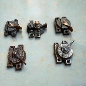 May include: An assortment of antique metal latches in various designs. The latches feature ornate details, including floral patterns and lever handles. The metal has a tarnished, aged appearance, with shades of bronze and silver.