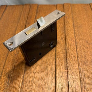 May include: A vintage door lock with a black body and a silver-colored metal plate. The plate has a rectangular brass-colored insert and a silver-colored latch. The lock is mounted on a wooden surface.