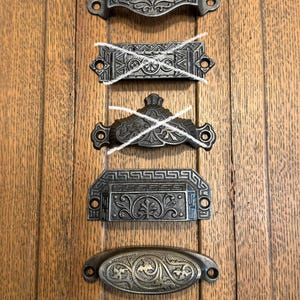 May include: A collection of five dark metal drawer pulls with ornate embossed patterns. Three of the pulls are rectangular with decorative borders and floral motifs, while one is oval-shaped with intricate scrollwork and one has a top slot.