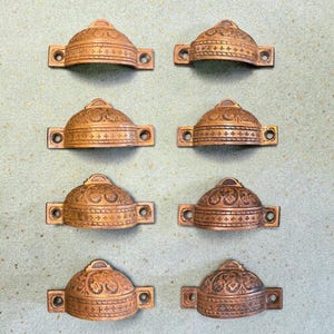 May include: Eight antique bronze cabinet pulls. Each pull has a domed top with ornate floral and cross designs. The pulls have rectangular mounting plates with two holes for screws. The pulls are approximately 4 inches wide.
