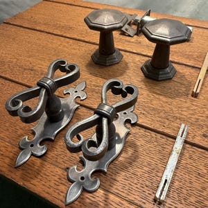 May include: A collection of antique door hardware, including two octagonal doorknobs, two ornate door handles with heart-shaped details, and several metal pieces. The hardware has a weathered, metallic finish, suggesting age and use. The items are arranged on a wooden surface.