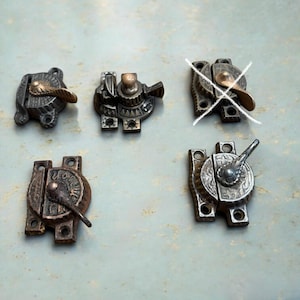 May include: Five metal window sash locks with varying patinas and decorative engravings. Each latch features a rotating lever mechanism and mounting plates with screw holes. One lock is marked with a white cross, indicating it is not available.