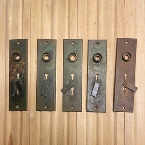 May include: Five antique bronze door plates with keyholes and lever-style latches. The plates are rectangular with a tarnished patina, showing signs of age and use. The background is a light-colored wooden surface.
