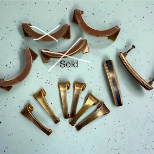 May include: A collection of metal cabinet hardware including three curved bronze-toned drawer pulls, one rectangular pull with a black center stripe, and five smaller angled metal handles, with the word Sold written over two items.