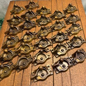 May include: A collection of antique brass cabinet latches. Each latch features a circular handle and a rectangular base with screw holes. The latches are arranged on a wooden surface, showcasing their aged patina and intricate design.