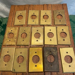 May include: A collection of rectangular brass electrical outlet covers. Each cover has a circular opening in the center and two smaller holes above and below. The brass has a variety of patinas, ranging from bright gold to dark brown.