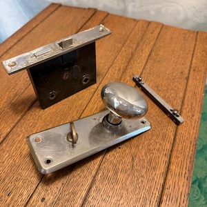 May include: An assortment of vintage door hardware, including a rectangular black lock mechanism, a silver door handle with an oval knob, and a small silver latch. The items are arranged on a wooden surface with visible grain.