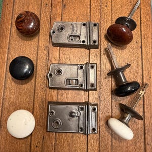 May include: A collection of vintage door hardware, including three metal latches and various doorknobs. The doorknobs are in shades of brown, black, and white. The hardware is arranged on a wooden surface, showcasing the antique style.