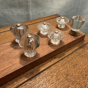 May include: A collection of clear glass cabinet knobs with various designs. Some knobs have a faceted, geometric shape, while others are round with a decorative top. The knobs are displayed on a wooden surface, showcasing their vintage aesthetic.