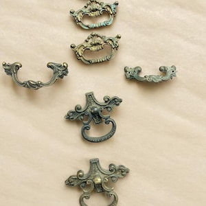May include: An assortment of antique brass drawer pulls with ornate designs. The pulls feature a mix of gold and verdigris finishes, showcasing intricate details and varying shapes. These vintage hardware pieces are ideal for furniture restoration projects.