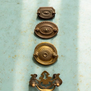 May include: Four antique brass drawer pulls in various designs. The top pull is rectangular, the second is oval, the third is round with a spiral design, and the bottom pull is a decorative handle with ornate details.