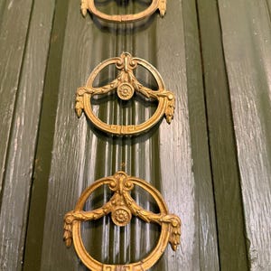 May include: Three antique brass door pulls with a circular design and ornate floral details are mounted vertically on a green wooden door. The pulls have a gold finish and are evenly spaced.