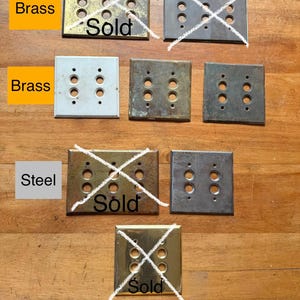 May include: A collection of rectangular metal wall plates for light switches, featuring various finishes including brass and steel. Several plates are marked with the word Sold and crossed out with white lines. The plates have multiple circular cutouts.