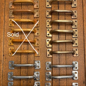 May include: A collection of vintage door pulls in various finishes. The image shows several rows of pulls in brass, bronze, and chrome. One set of pulls is marked "Sold". The pulls have a rectangular base with rounded handles.