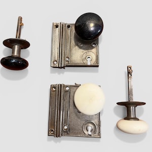 May include: Antique door hardware set featuring two door knobs and two mortise locks. One set has a black knob, the other a white knob. The locks are rectangular with decorative vertical lines and keyholes.