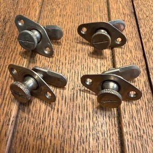 May include: A collection of vintage metal window latches. Each latch features a screw-down knob, a pivoting lever, and a rectangular base with two screw holes. The latches are made of a silver-toned metal and are arranged on a wooden surface.