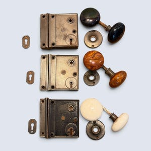 May include: Three sets of rectangular metal rim locks with matching keyhole escutcheons and door knobs. The knobs feature distinct finishes: one black, one brown wood-grain pattern, and one white ceramic. Each lock includes a square spindle hole.