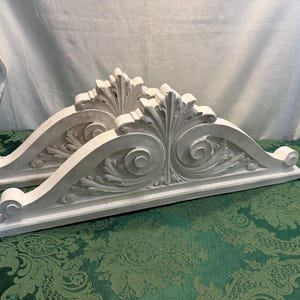 May include: Two white architectural corbels featuring intricate carved scrollwork and acanthus leaf motifs. Each piece has a flat base, a curved top edge, and a central vertical leaf design, showcasing a symmetrical pattern on both sides.