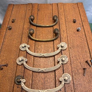 May include: Four sets of antique drawer pulls and hardware. Two sets are brass-toned, curved handles. The other two sets are off-white, ornate pulls with decorative backplates. All are mounted on a wooden surface with screws.