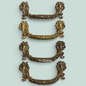 May include: Four antique brass drawer pulls. Each pull has ornate, detailed end pieces with a floral design and a textured, curved handle. The brass has a warm, aged patina, suggesting vintage or antique hardware for furniture restoration.