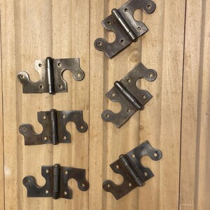 May include: Six antique metal hinges with a dark, aged finish. Each hinge has a decorative shape with rounded edges and multiple screw holes. The hinges are arranged on a light-colored wooden surface.